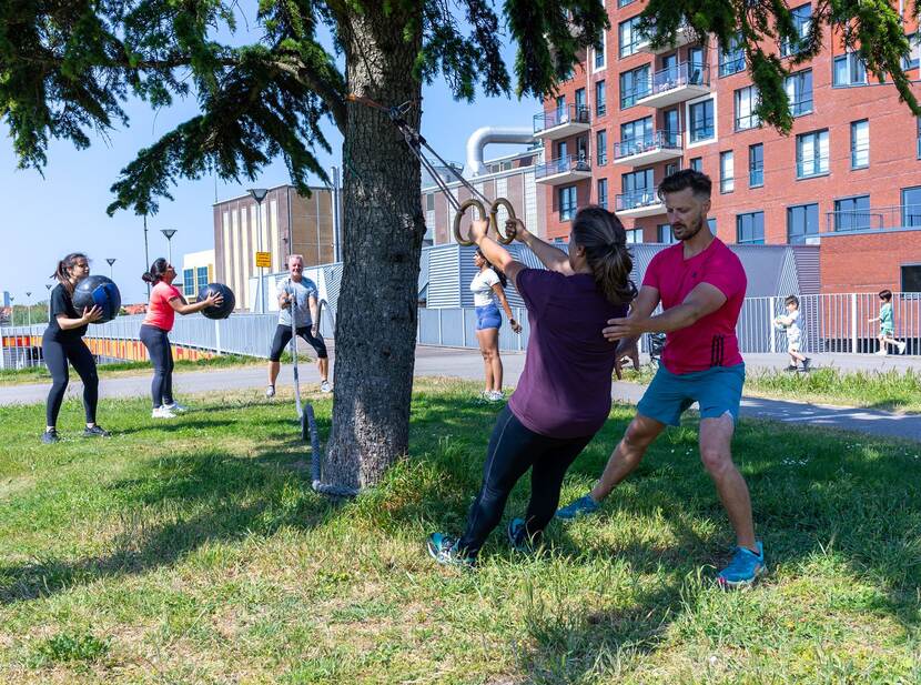 Op de foto is een groep mensen buiten aan het sporten. Vooraan staat een vrouw die wordt geholpen door de instructeur.