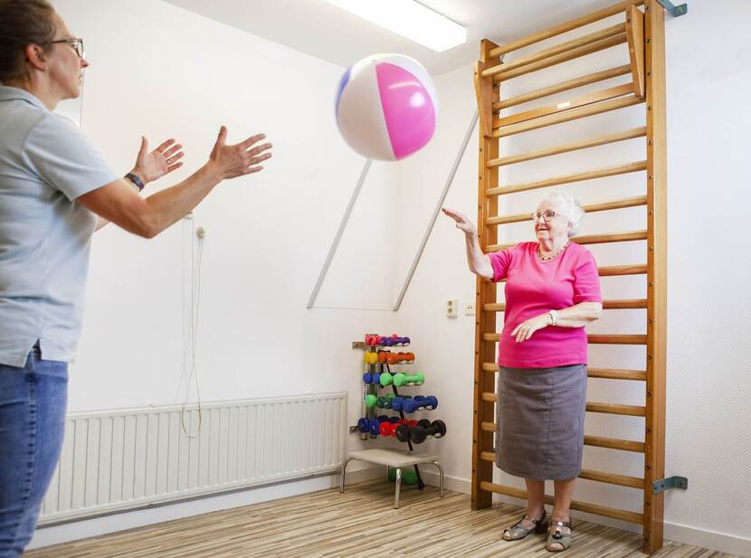 De foto toont een oudere dame die als oefening een bal overgooit met een oefentherapeut.
